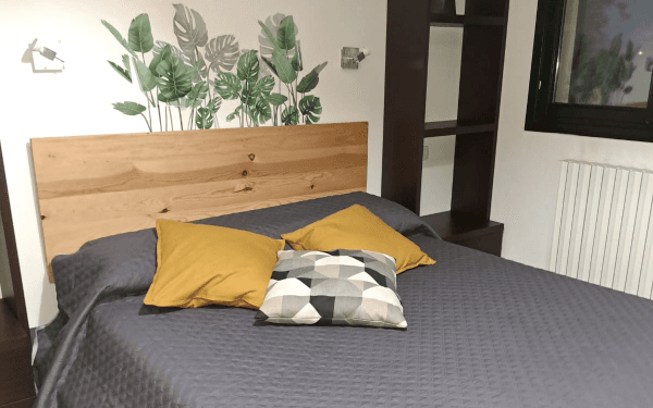 Dettaglio del letto matrimoniale con copriletto grigio, cuscini gialli e decorazione murale a foglie